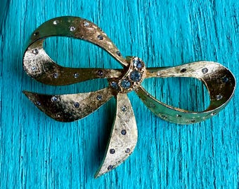 May include: A gold-toned bow brooch with a textured surface. The bow is adorned with small, clear rhinestones and a cluster of blue rhinestones at the center. The brooch has a classic, elegant design.