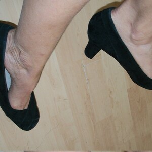 Peut inclure: Une paire de chaussures &agrave; talons en daim noir avec une pointe pointue. Les chaussures sont port&eacute;es sur un sol en bois.