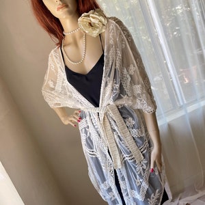 May include: A mannequin wearing a cream-colored, floral lace kimono with a matching belt. The kimono is over a black top and shorts. A pearl necklace and a large cream-colored flower are also visible.