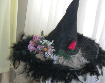 May include: A black pointed hat with a wide brim adorned with black feathers, a cluster of purple and white artificial flowers, and a single red rose. The hat is displayed on a white metal stand with a tripod base and curved legs.