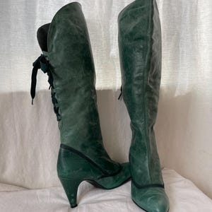 May include: A pair of green leather knee-high boots with a pointed toe and a stiletto heel. The boots have a lace-up detail on the back of the shaft.