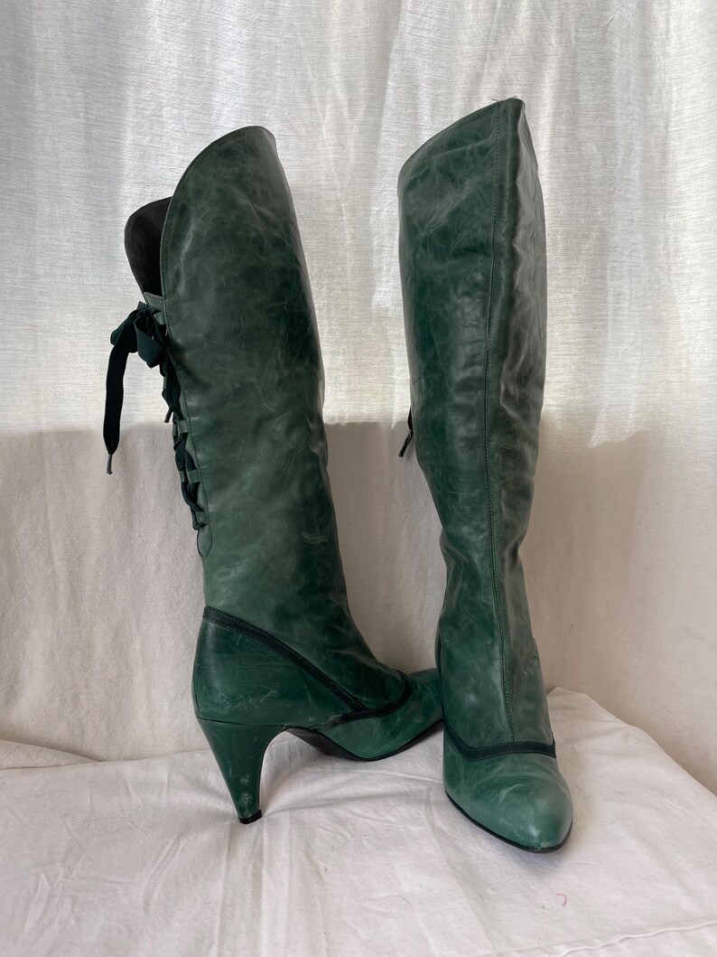May include: A pair of green leather knee-high boots with a pointed toe and a stiletto heel. The boots have a lace-up detail on the back of the shaft.