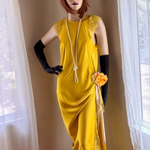May include: A mustard yellow satin dress with lace accents and a draped skirt. The dress is accessorized with a pearl necklace, black gloves, and a floral embellishment. A black and gold headband with feathers completes the look.