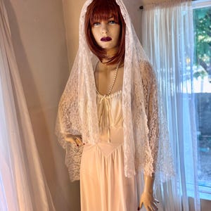May include: A vintage-style, ivory-colored bridal ensemble on a mannequin. The outfit includes a long, flowing gown and a delicate lace veil. A pearl necklace adorns the mannequin's neck. The veil is draped over the head and shoulders, creating a romantic look.