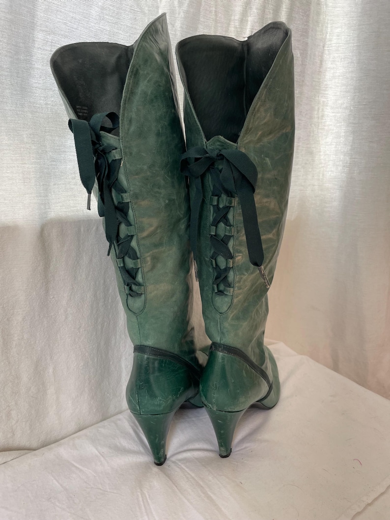 May include: A pair of green leather knee-high boots with black laces up the back. The boots have a pointed toe and a high heel.
