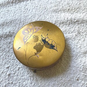 May include: A round, gold-colored compact with a floral design featuring butterflies in pink, blue, and white.