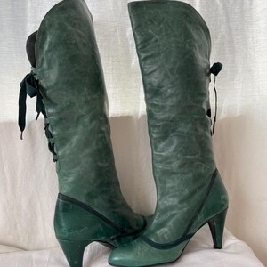 May include: A pair of green leather knee-high boots with a pointed toe and a high heel. The boots have a lace-up detail on the back of the shaft.