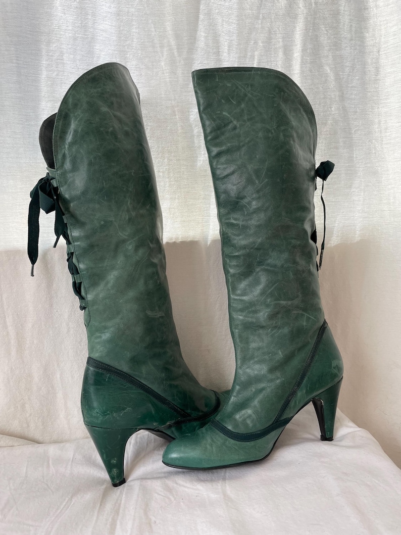 May include: A pair of green leather knee-high boots with a pointed toe and a high heel. The boots have a lace-up detail on the back of the shaft.