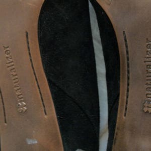 Peut inclure: Gros plan sur le dessous de deux chaussures en daim noir avec des semelles int&eacute;rieures en cuir marron. Les semelles int&eacute;rieures portent le texte "Naturalizer" imprim&eacute;.