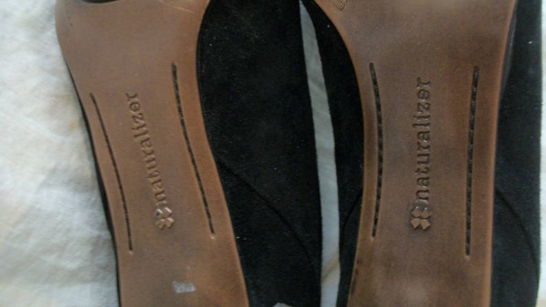 Peut inclure: Gros plan sur le dessous de deux chaussures en daim noir avec des semelles int&eacute;rieures en cuir marron. Les semelles int&eacute;rieures portent le texte "Naturalizer" imprim&eacute;.