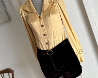 May include: A yellow satin blouse with a collared neckline and long sleeves. The blouse has a button-up front and a fitted silhouette. The blouse is paired with black velvet shorts with a silver belt.