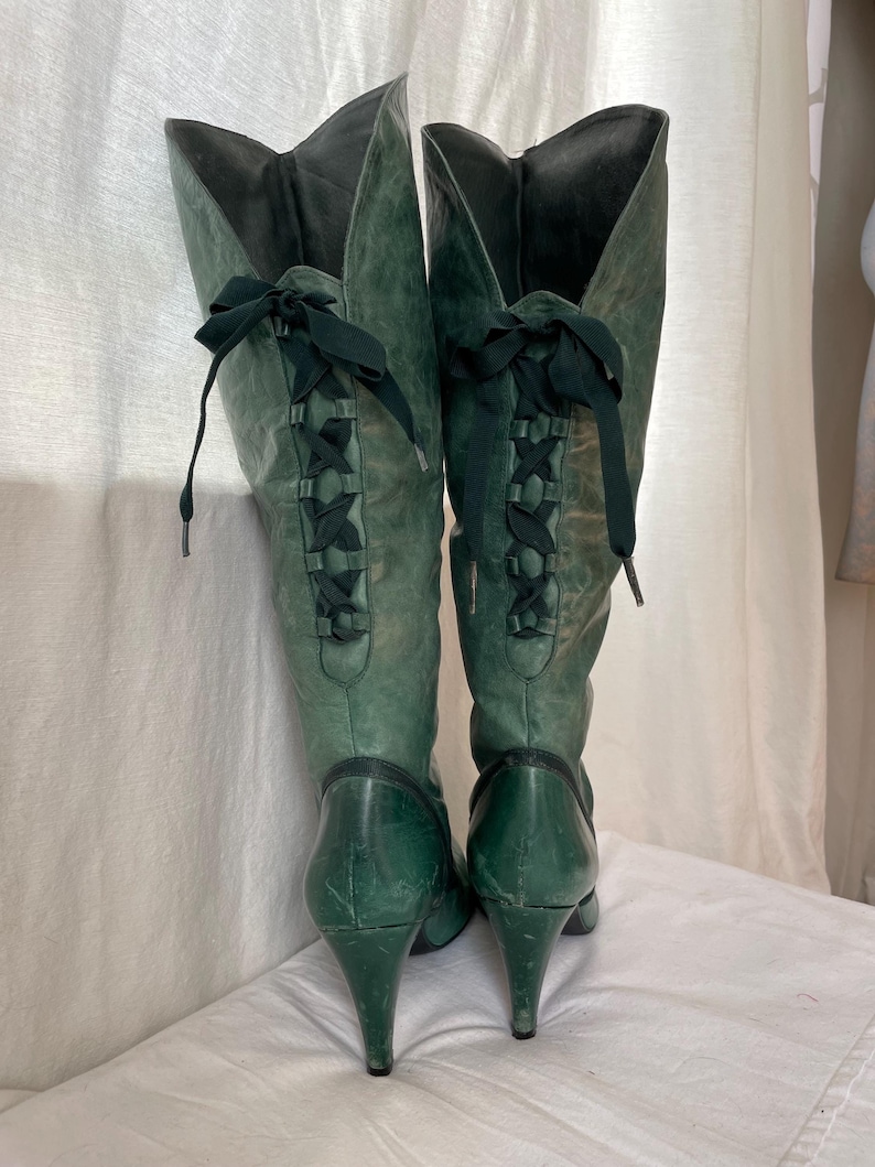 May include: A pair of green leather knee-high boots with black ribbon laces up the back. The boots have a pointed toe and a high heel.