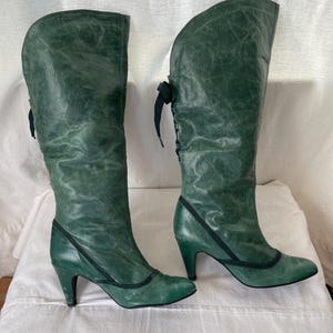 May include: A pair of green leather knee-high boots with a lace-up detail on the back of the boot. The boots have a high heel and a pointed toe.