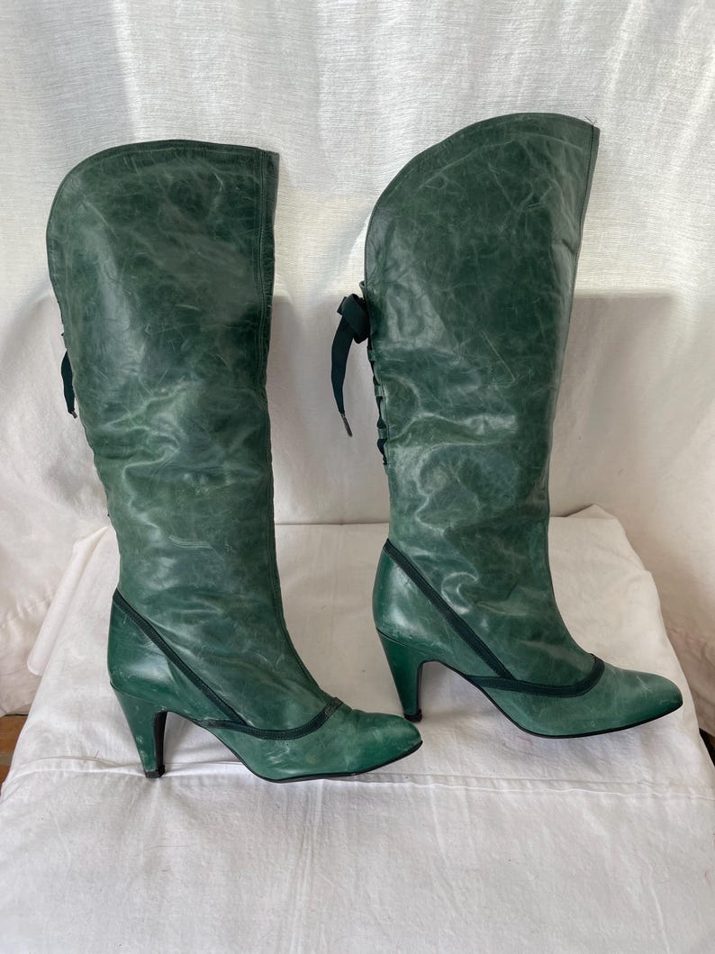 May include: A pair of green leather knee-high boots with a lace-up detail on the back of the boot. The boots have a high heel and a pointed toe.