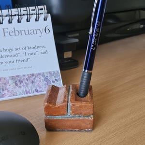 May include: A blue pen with silver accents is in a brick-shaped pen holder. The pen has the text "TW Kempton (C.C.) Ltd Tel +44(01116 261 0001" engraved on it.