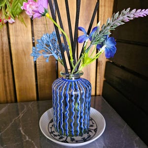 May include: A glass diffuser bottle with blue and white vertical stripes, containing black reeds and artificial flowers. The bottle rests on a white plate with a black floral pattern. The background is a wooden wall.