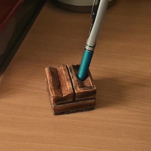 May include: A wooden pen holder with a rustic design. The holder has two slots for pens and is made of brown wood with a dark brown stain. A blue and silver pen is in the holder.