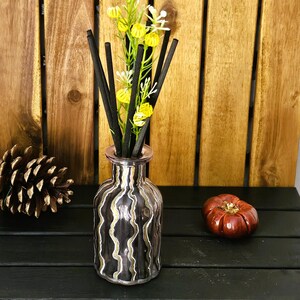 May include: A black and gold striped glass bottle with black diffuser sticks and yellow flowers. The bottle is sitting on a black wooden surface.