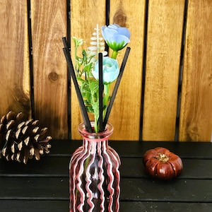 May include: A pink glass diffuser bottle with white wavy lines and black diffuser sticks. The bottle is filled with a clear liquid and has a bouquet of blue and white flowers on top.