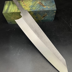 May include: A long, thin, silver knife blade with a textured finish. The blade is resting on a block of green and blue wood.