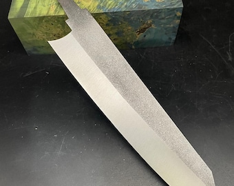 May include: A long, thin, silver knife blade with a textured finish. The blade is resting on a block of green and blue wood.