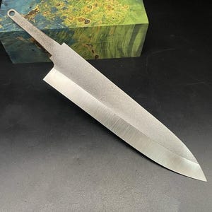 Könnte beinhalten: Eine silberne Messerklinge mit strukturierter Oberfläche und einem Griff mit einem Loch oben. Die Klinge ist scharf und spitz. Das Messer liegt auf einer schwarzen Oberfläche.