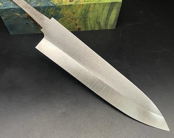 May include: A silver knife blade with a textured finish and a handle with a hole at the top. The blade is sharp and pointed. The knife is on a black surface.