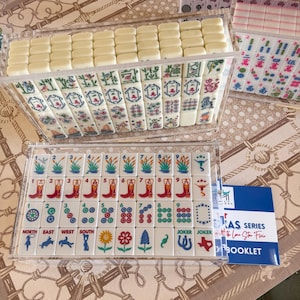 May include: A set of Mahjong tiles in clear acrylic cases, featuring white tiles with colorful illustrations of cowboy boots, Texas maps, flowers, and western motifs. A blue and white booklet labeled Texas Series with the Lone Star Flair is included.