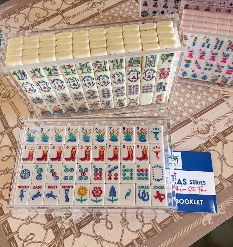 May include: A set of Mahjong tiles in clear acrylic cases, featuring white tiles with colorful illustrations of cowboy boots, Texas maps, flowers, and western motifs. A blue and white booklet labeled Texas Series with the Lone Star Flair is included.