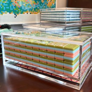 May include: A set of rectangular mahjong tiles stacked in a clear acrylic display case. Each tile features a three-layered design with an olive green top, a light blue middle, and a peach-colored base. The tiles are arranged in a grid pattern.