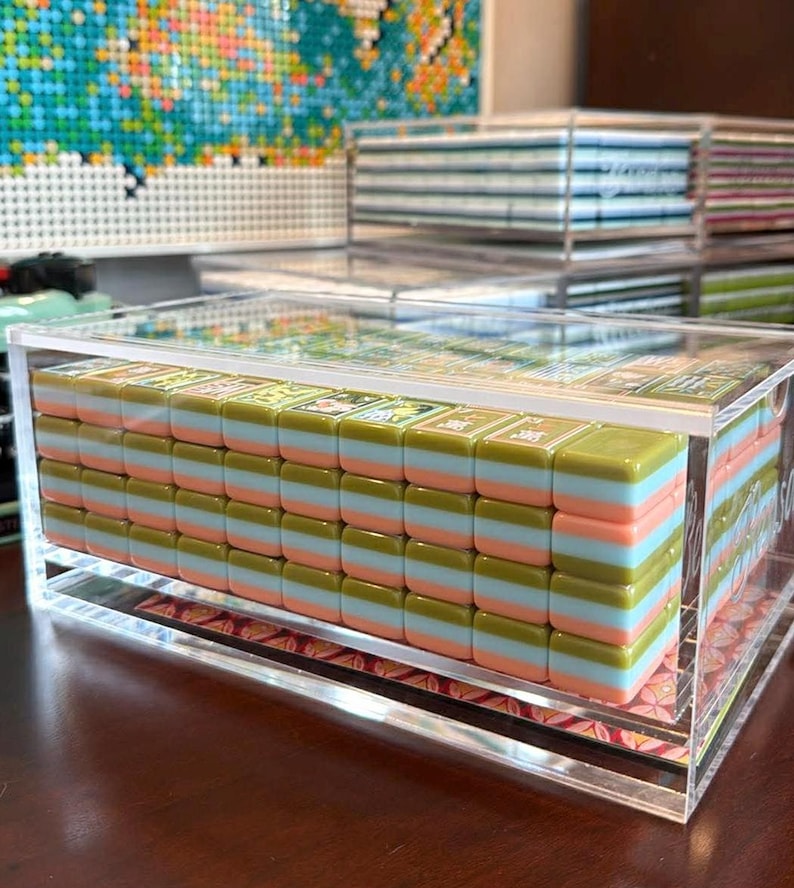 May include: A set of rectangular mahjong tiles stacked in a clear acrylic display case. Each tile features a three-layered design with an olive green top, a light blue middle, and a peach-colored base. The tiles are arranged in a grid pattern.