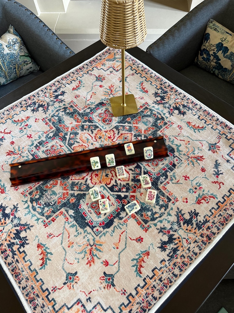 May include: A square tablecloth with a cream base and a multi-colored floral and geometric pattern in shades of navy blue, orange, and teal. A long, dark brown tortoiseshell-patterned mahjong tile rack holds several tiles with colorful designs.