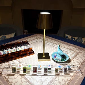 May include: A gold-toned metal table lamp with a conical shade stands on a table alongside a clear acrylic tile rack, a tortoiseshell-patterned tile rack, and a small blue decorative dish containing several colorful dice and a bird figurine.