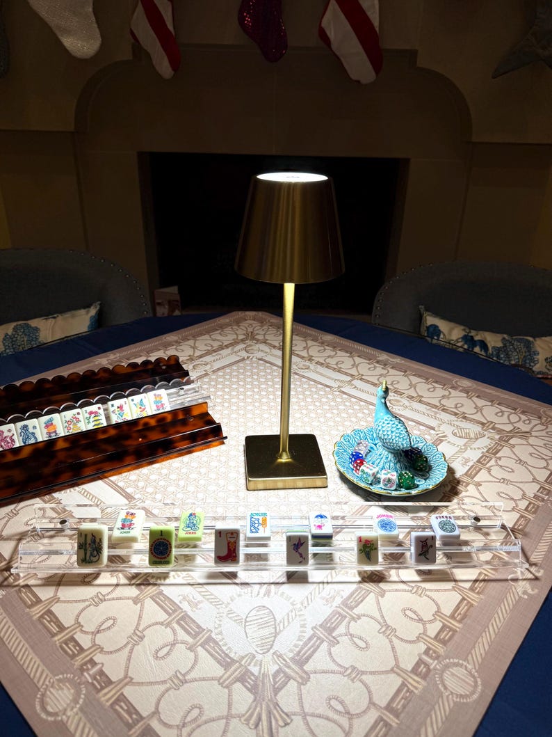 May include: A gold-toned metal table lamp with a conical shade stands on a table alongside a clear acrylic tile rack, a tortoiseshell-patterned tile rack, and a small blue decorative dish containing several colorful dice and a bird figurine.