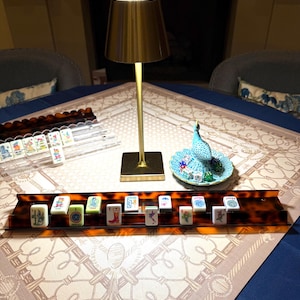 May include: A mahjong set featuring white tiles with colorful illustrations, arranged on a brown tortoiseshell-patterned rack. A gold-toned metal table lamp stands nearby, alongside a small blue ceramic peacock dish holding several dice.