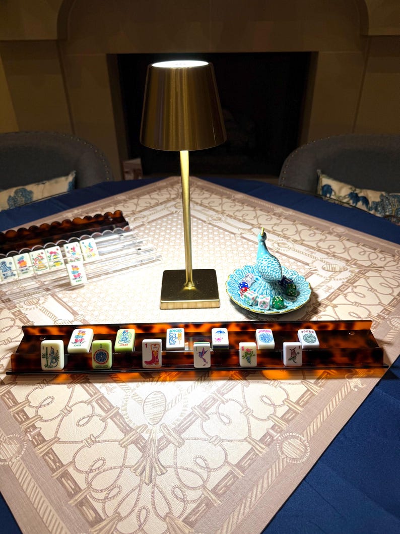 May include: A mahjong set featuring white tiles with colorful illustrations, arranged on a brown tortoiseshell-patterned rack. A gold-toned metal table lamp stands nearby, alongside a small blue ceramic peacock dish holding several dice.