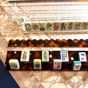 May include: A set of Mahjong tiles featuring colorful illustrations of animals, objects, and symbols, arranged on two tiered racks. The top rack is clear acrylic, while the bottom rack is a brown tortoiseshell pattern with a scalloped edge.