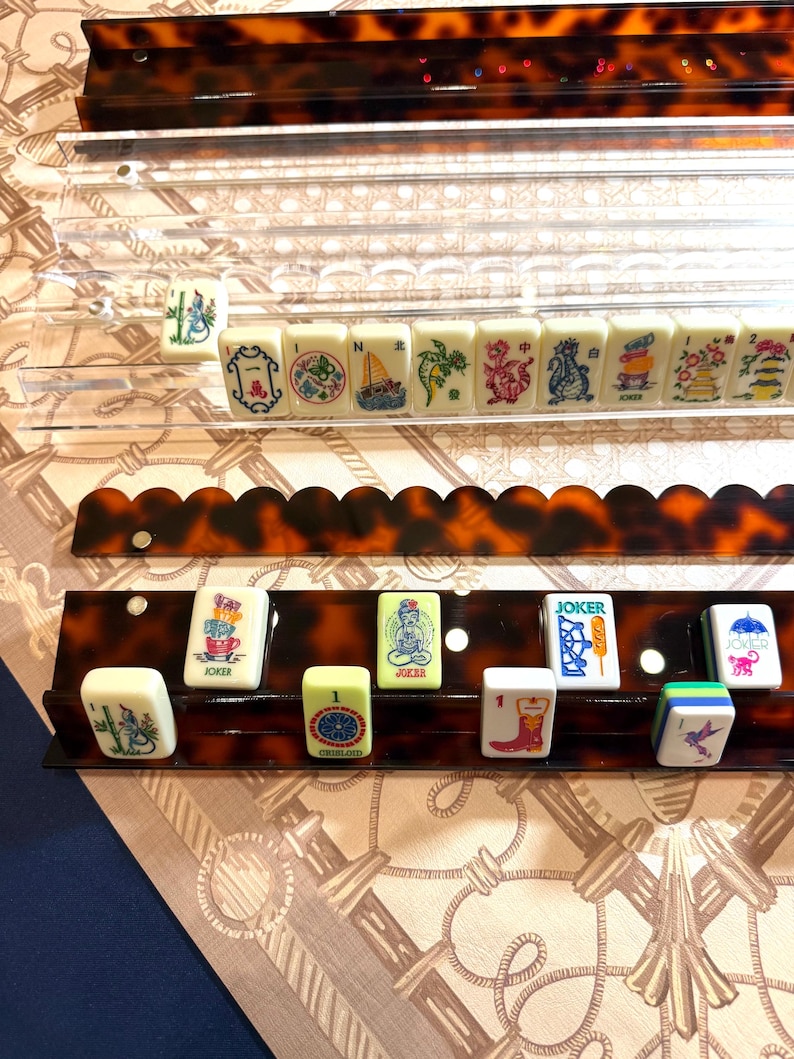 May include: A set of Mahjong tiles featuring colorful illustrations of animals, objects, and symbols, arranged on two tiered racks. The top rack is clear acrylic, while the bottom rack is a brown tortoiseshell pattern with a scalloped edge.