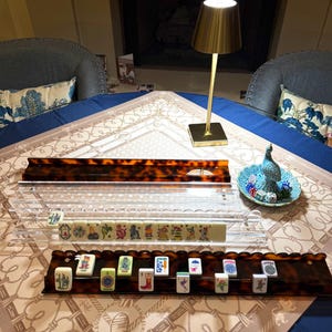 May include: A mahjong set featuring two tortoiseshell-patterned racks, one clear acrylic rack, and a small teal dish containing dice. The racks hold various ivory-colored tiles with colorful graphic illustrations, including a joker tile.