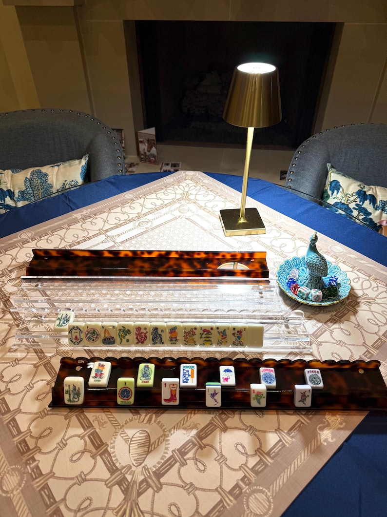 May include: A mahjong set featuring two tortoiseshell-patterned racks, one clear acrylic rack, and a small teal dish containing dice. The racks hold various ivory-colored tiles with colorful graphic illustrations, including a joker tile.