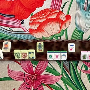 May include: A brown tortoiseshell patterned Mahjong tile rack holding a row of rectangular white tiles. The tiles feature various colorful engraved illustrations, including dragons, flowers, and the word Joker, arranged horizontally on the rack.