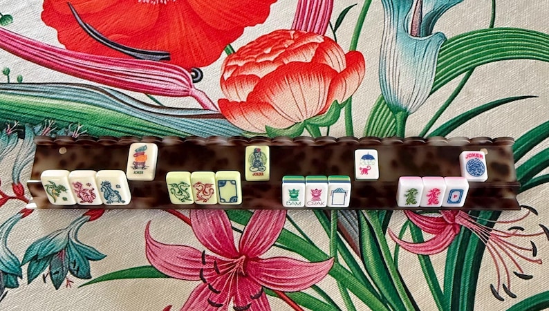 May include: A brown tortoiseshell patterned Mahjong tile rack holding a row of rectangular white tiles. The tiles feature various colorful engraved illustrations, including dragons, flowers, and the word Joker, arranged horizontally on the rack.