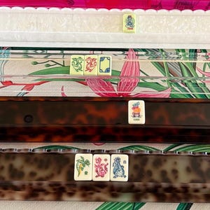 May include: Four mahjong tile racks arranged horizontally. The top rack is white with a scalloped edge, the second is clear acrylic, the third is dark brown tortoiseshell, and the bottom is a lighter brown tortoiseshell. Each rack holds mahjong tiles.