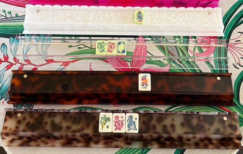 May include: Four mahjong tile racks arranged horizontally. The top rack is white with a scalloped edge, the second is clear acrylic, the third is dark brown tortoiseshell, and the bottom is a lighter brown tortoiseshell. Each rack holds mahjong tiles.