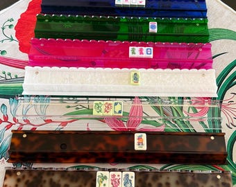 Puede incluir: Un conjunto de siete soportes rectangulares para fichas de Mahjong en varios colores y acabados, incluyendo azul, verde, rosa, transparente, blanco translúcido y dos patrones de carey marrón, cada uno con una pequeña selección de fichas ilustradas.