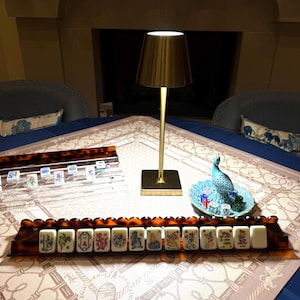 May include: A mahjong set featuring two tortoiseshell-patterned racks holding white tiles with colorful illustrations, a gold-toned metal table lamp with a conical shade, and a small blue decorative dish containing several translucent dice.