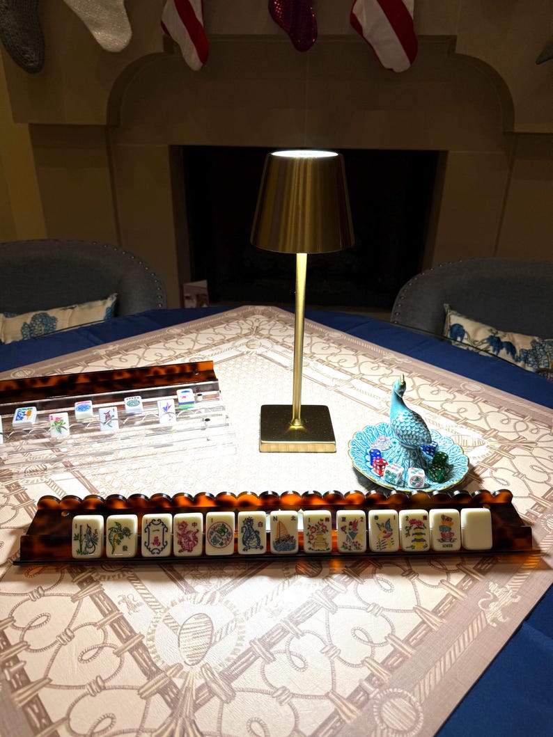 May include: A mahjong set featuring two tortoiseshell-patterned racks holding white tiles with colorful illustrations, a gold-toned metal table lamp with a conical shade, and a small blue decorative dish containing several translucent dice.