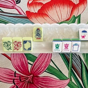 May include: A white plastic mahjong tile rack holding a row of rectangular tiles. The tiles feature colorful illustrations of dragons, floral patterns, and text including "JOKER", "BAM", and "CRAK". The rack has a scalloped top edge and a textured base.