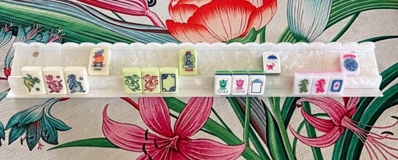 May include: A white plastic mahjong tile rack holding a row of rectangular tiles. The tiles feature colorful illustrations of dragons, floral patterns, and text including "JOKER", "BAM", and "CRAK". The rack has a scalloped top edge and a textured base.
