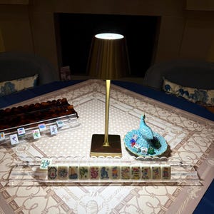 May include: A gold-toned metal table lamp with a conical shade stands next to a clear acrylic rack holding mahjong tiles. A small turquoise ceramic peacock figurine sits on a matching scalloped dish containing several colorful dice.